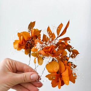 May include: A set of three hair pins adorned with dried orange flowers and white baby's breath. The flowers are arranged in a cluster and secured to the pins with wire.