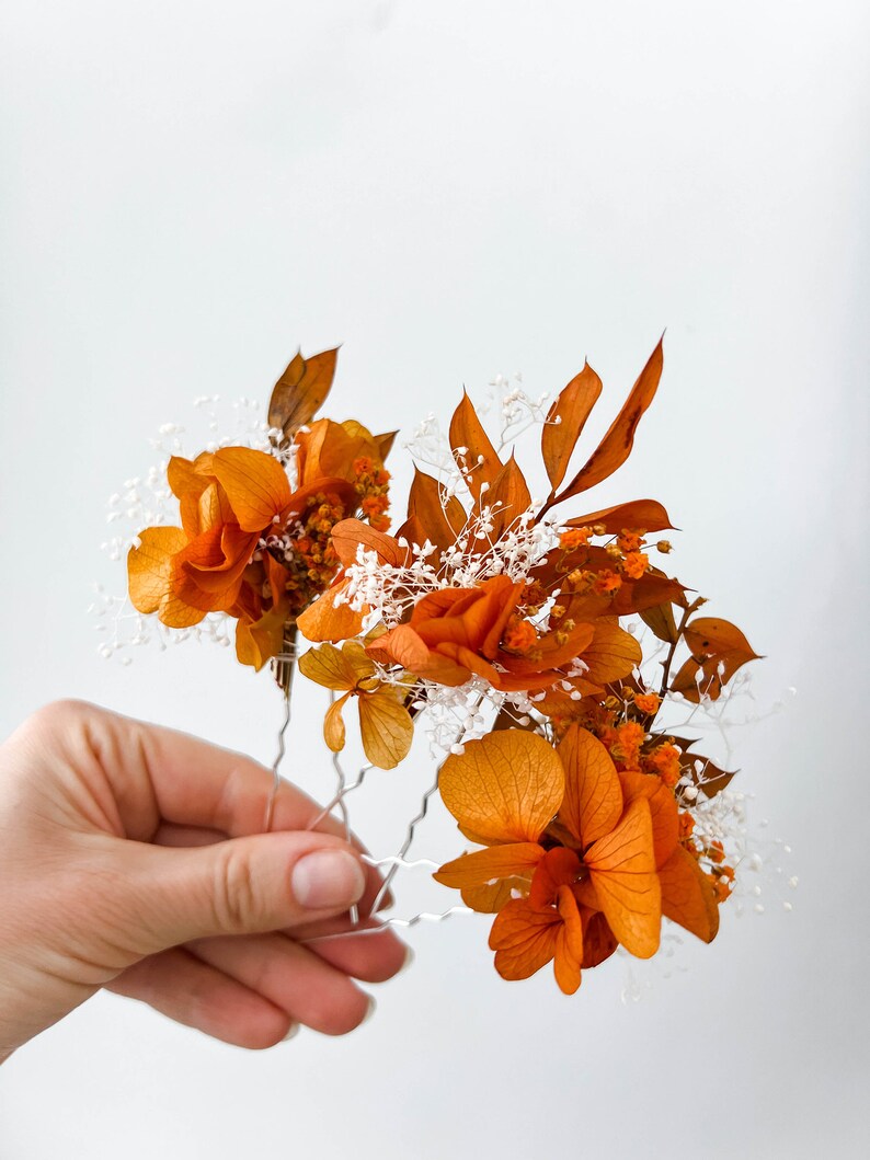 May include: A set of three hair pins adorned with dried orange flowers and white baby's breath. The flowers are arranged in a cluster and secured to the pins with wire.