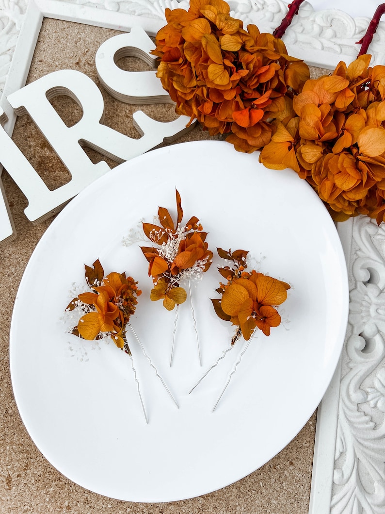 May include: Three hair pins with dried orange flowers and white sprigs. The pins are silver and have a wavy design.