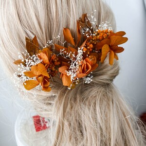May include: Three dried flower hair clips with orange and white flowers and greenery. The clips are arranged in a row on a blonde wig.