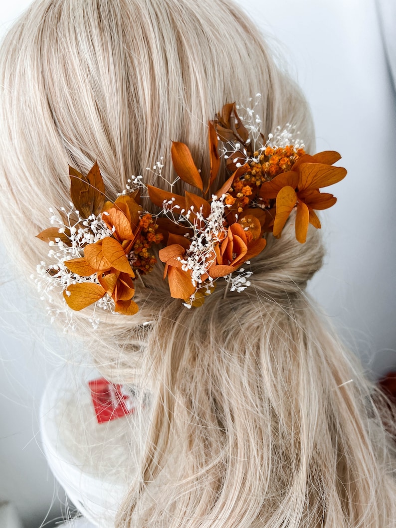 May include: Three dried flower hair clips with orange and white flowers and greenery. The clips are arranged in a row on a blonde wig.
