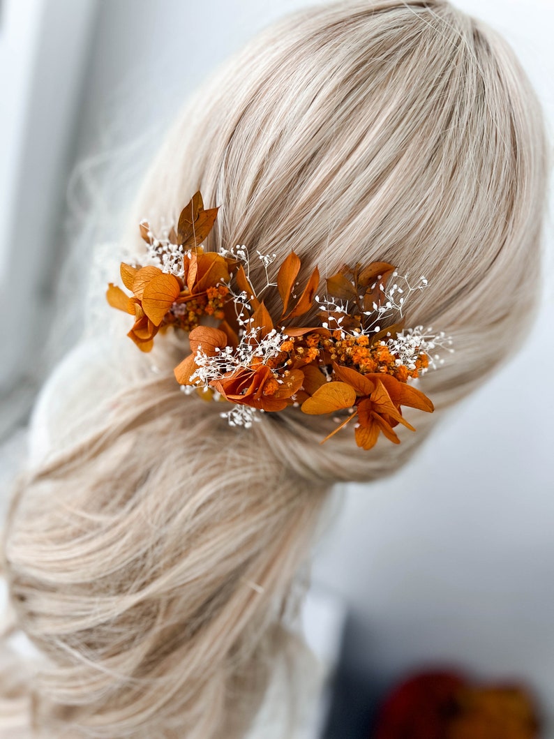 May include: A hair accessory with dried orange flowers and white baby's breath. The flowers are arranged in a cluster and are attached to a hair clip.