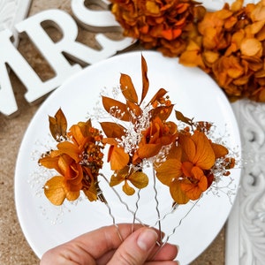 May include: Four hair clips with dried orange flowers and white baby's breath. The clips are silver and have a wavy design.