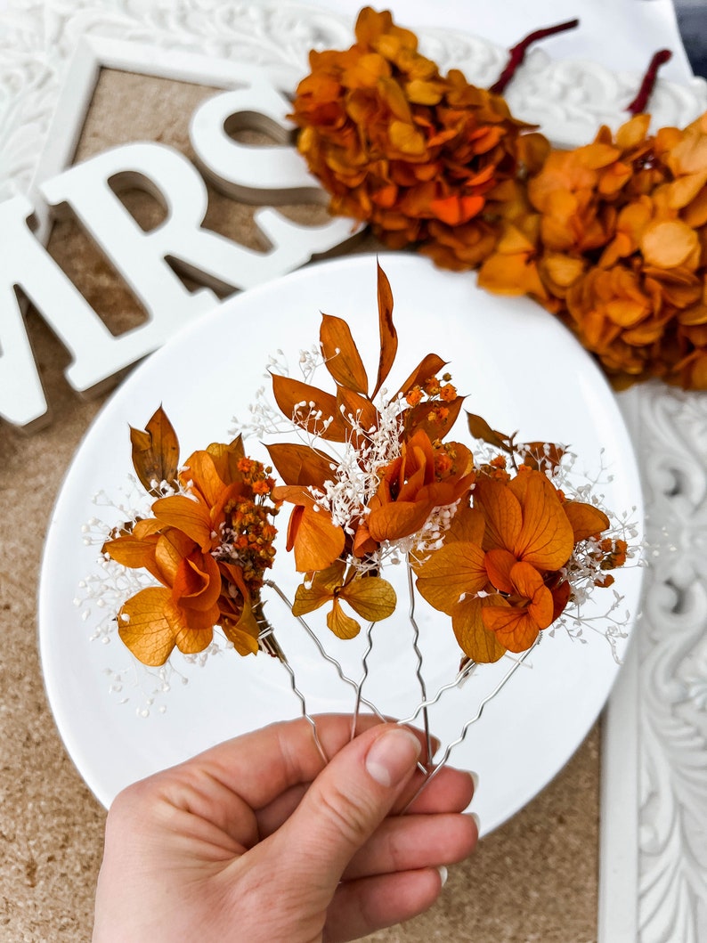 May include: Four hair clips with dried orange flowers and white baby's breath. The clips are silver and have a wavy design.