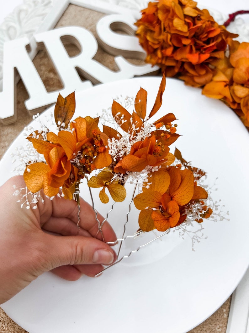 May include: Three hair clips with dried orange flowers and white baby's breath. The clips are silver and have a wavy design.