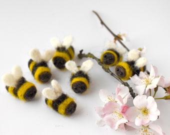 May include: A group of seven felt bee ornaments with black and yellow stripes and white wings. The bees are arranged on a white surface with a branch of pink blossoms.