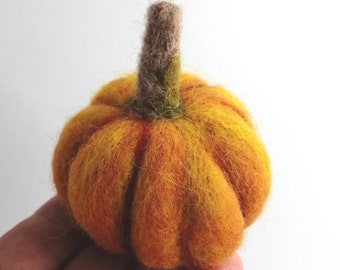 May include: A small, round, needle-felted pumpkin crafted from soft wool fibers in vibrant shades of orange and yellow. The pumpkin features a textured surface with vertical ridges and a short, upright stem in a muted brown and green color.