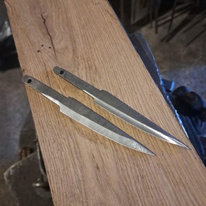 May include: Two unfinished metal knife blades with rounded tips, lying on a wooden surface.