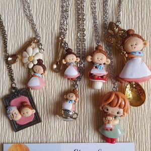 Puede incluir: Una colección de collares hechos a mano de arcilla polimérica con figuras en miniatura de niñas que llevan vestidos y sostienen varios accesorios. Los collares se muestran sobre un fondo marrón claro con una tarjeta de visita para "Elvira Carangi" con el texto "creazioni modellate a mano in pasta polimerica handmade polymer clay creations" e información de contacto.