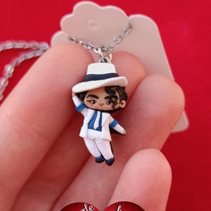 Op de afbeelding: Een zilverkleurige ketting met een kleine, geboetseerde polymeerklei-figuur van een persoon in een wit pak, blauw overhemd en witte fedora-hoed. De figuur is afgebeeld met één hand die de rand van de hoed aanraakt.
