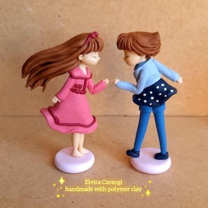 May include: Two polymer clay figurines of people, one in a pink dress and the other in a blue jacket and skirt with white polka dots. The figurines are on round, light pink bases. Text reads "Elvira Carangi handmade with polymer clay."
