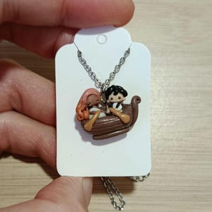 May include: A silver necklace with a pendant depicting a brown boat with two small figures. One figure has pink hair, and the other has black hair. The pendant is suspended from a silver chain.