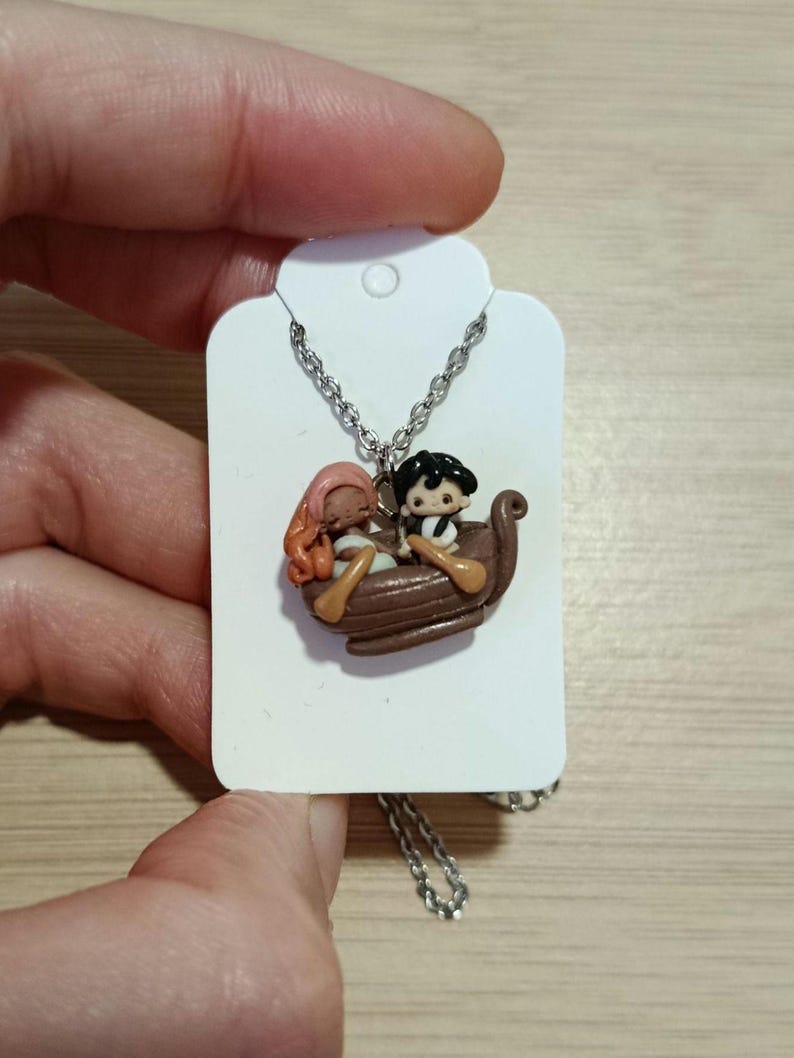 May include: A silver necklace with a pendant depicting a brown boat with two small figures. One figure has pink hair, and the other has black hair. The pendant is suspended from a silver chain.