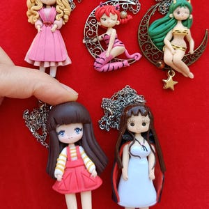Könnte beinhalten: Fünf Halsketten mit modellierten Figuren. Eine mit blondem Haar in einem rosa Kleid, eine rothaarige Figur auf einer Mondsichel, eine grünhaarige Figur auf einem Mond mit Stern, eine mit braunem Haar in rotem Outfit und eine in einem weißen Kleid.