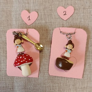 Può includere: Due collane con ciondolo fatte a mano su biglietti rosa. Una presenta una figura in miniatura su un fungo rosso e bianco, con un cucchiaio d'oro. L'altra ha una figura su un oggetto marrone. Entrambe hanno catene d'argento.
