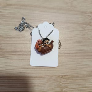 May include: A silver necklace with a pendant depicting a brown boat with two cartoon figures. One figure has black hair and the other has red hair. The pendant is on a silver chain and is displayed on a white tag.