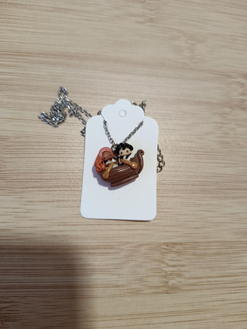 May include: A silver necklace with a pendant depicting a brown boat with two cartoon figures. One figure has black hair and the other has red hair. The pendant is on a silver chain and is displayed on a white tag.