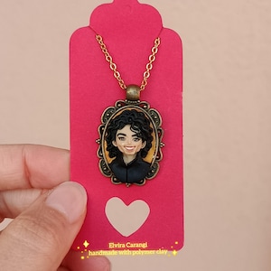 Könnte beinhalten: Eine handgefertigte Polymer Clay Anhänger Halskette mit einem Porträt einer Person mit dunklem, lockigem Haar und einer schwarzen Jacke. Das Porträt befindet sich in einem ovalen, verzierten Rahmen mit einer goldfarbenen Kette. Der Anhänger hängt an einem rosa Anhänger.