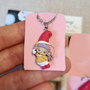 Könnte beinhalten: Eine handgefertigte Halskette mit einer Miniaturfigur eines Mädchens mit roter Weihnachtsmütze und gelbem Kleid. Der Anhänger ist an einer silbernen Kette befestigt und auf einer rosa Karte präsentiert. Die Figur ist etwa 2,5 cm groß.