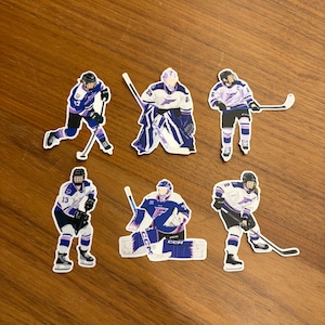 May include: A collection of six die-cut stickers featuring hockey players in action. The stickers depict players in white, purple, and blue uniforms, with hockey sticks and protective gear. The stickers are on a wooden surface.
