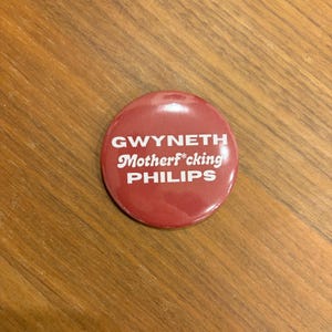 May include: A circular, red button with the white text "GWYNETH Motherf*cking PHILIPS". The button is placed on a wooden surface.