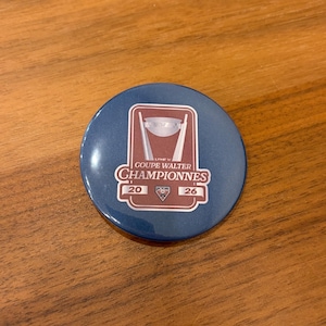 Può includere: Un bottone rotondo blu con la scritta "COUPE WALTER CHAMPIONNES 2026" in bianco e rosso. Il bottone presenta una grafica stilizzata del trofeo. Il bottone è su una superficie di legno.