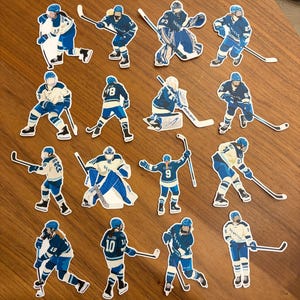 May include: A collection of die-cut stickers featuring hockey players in action. The stickers showcase players in blue and white uniforms, with various poses including skating, shooting, and goaltending. Each sticker has a unique player number.