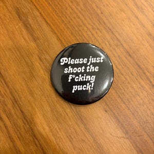 Può includere: Un bottone nero con testo bianco che recita "Please just shoot the f*cking puck!". Il bottone è rotondo e si trova su una superficie di legno.
