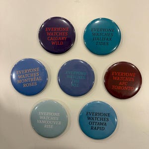 Può includere: Una collezione di sette bottoni rotondi in vari colori, ognuno con la frase "EVERYONE WATCHES" seguita da una città e dal nome di una squadra in testo bianco. I bottoni sono in tonalità di blu, verde acqua e bordeaux. Per squadre sportive.