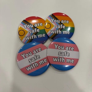 Puede incluir: Cuatro chapas redondas con el texto "You are safe with me" en letras negras. Dos chapas muestran la bandera Progress Pride con un círculo amarillo y un anillo morado, mientras que dos chapas muestran los colores de la bandera transgénero.
