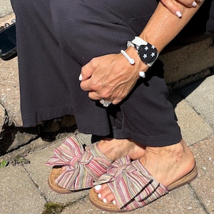 May include: A pair of multi-colored striped fabric slides with a large bow on the top of each shoe. The shoes are on a light brown cork platform.
