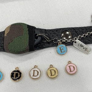 Puede incluir: Un brazalete de cuero negro con un inserto de tela de camuflaje y un dije plateado que dice "LOVE". El brazalete tiene una cadena dorada con un dije de letra "E" de esmalte azul y cinco dijes dorados con dijes de letra "D" de esmalte blanco, negro y azul.