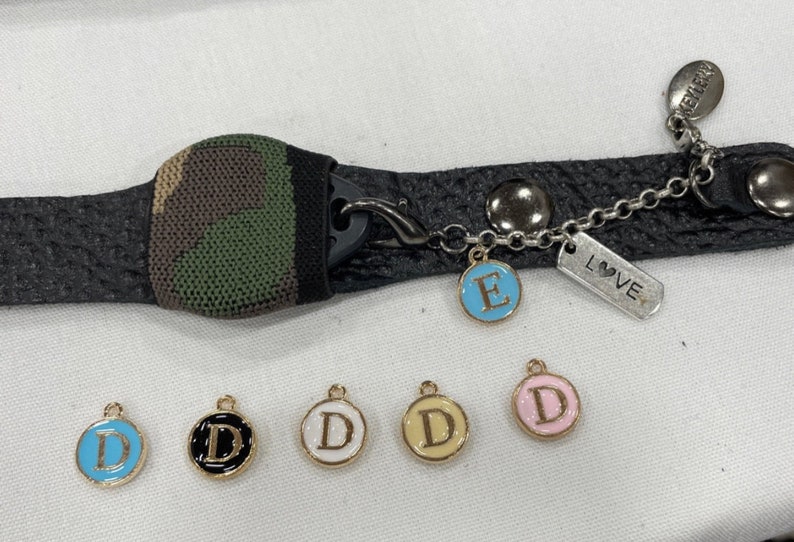 Puede incluir: Un brazalete de cuero negro con un inserto de tela de camuflaje y un dije plateado que dice "LOVE". El brazalete tiene una cadena dorada con un dije de letra "E" de esmalte azul y cinco dijes dorados con dijes de letra "D" de esmalte blanco, negro y azul.