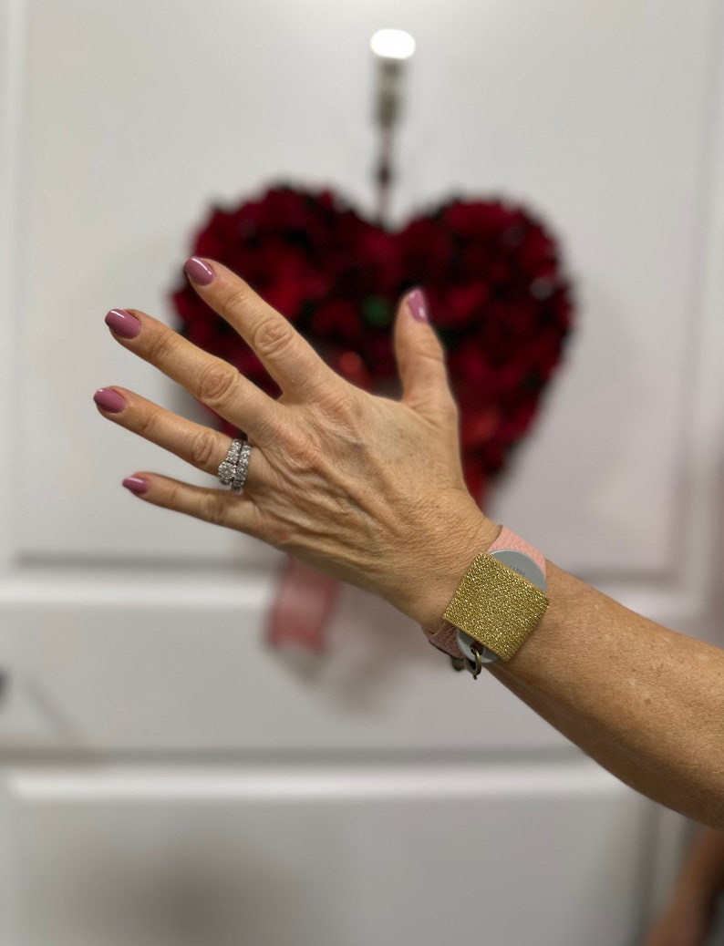 Puede incluir: Un brazalete de cuero rosa con un dije cuadrado dorado. El dije tiene una superficie texturizada.