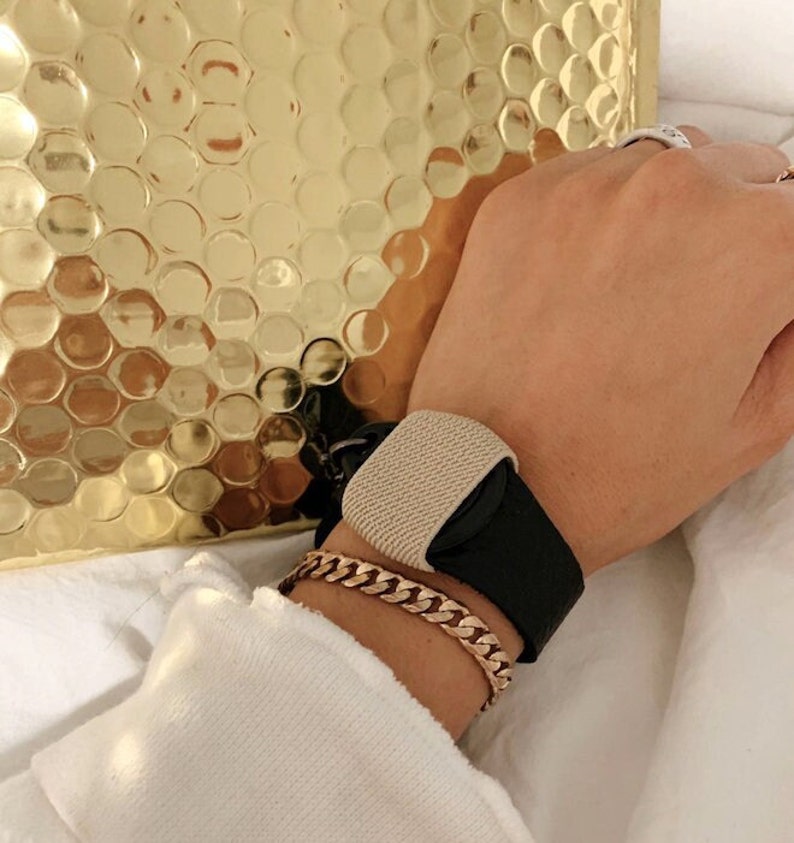 Puede incluir: Una pulsera negra y beige con una correa de tela texturizada y una hebilla negra. La pulsera se lleva en la mu&ntilde;eca de una persona.