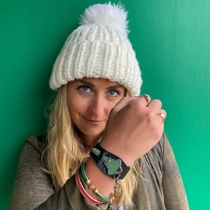 May include: A woman wearing a white knit beanie with a fluffy pom pom, a black leather bracelet with a green key charm, and several colorful bracelets on her wrist. She is wearing a brown jacket and has her hand covering her face.