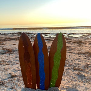 Könnte beinhalten: Drei Holzsurfboards mit bunten Harzmustern. Die Bretter stehen aufrecht im Sand an einem Strand. Die Bretter sind braun mit einem blauen, grünen und orangefarbenen Harzmuster.