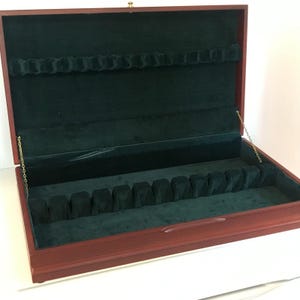 May include: A rectangular wooden storage chest with a dark reddish-brown finish, featuring a hinged lid held by two thin gold-colored chains. The interior is lined with dark green velvet and includes molded slots for organizing items.