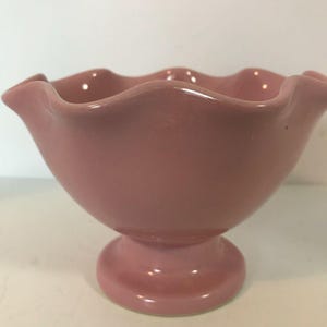 May include: A solid pink ceramic bowl with a smooth, glossy finish. The bowl features a wide, flared opening with a distinctively wavy, scalloped rim and is supported by a sturdy, circular pedestal base, creating a classic footed silhouette.