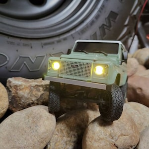 May include: A green toy Land Rover Defender driving over rocks. The toy has headlights and a grill.