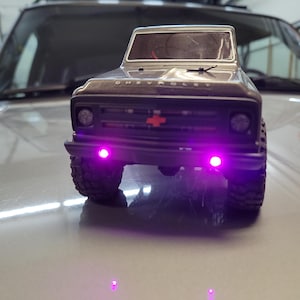 May include: A black and silver toy Chevrolet pickup truck with a red bowtie emblem on the front grill. The truck has purple LED lights on the front bumper and headlights. The tires are black and rugged.