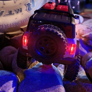 May include: A white toy truck with black tires and red taillights. The truck is driving over rocks and has a black roof rack with red containers on top.