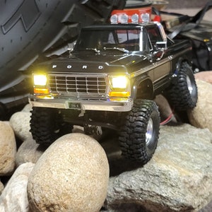 May include: Black Ford pickup truck with a silver grill and chrome bumpers. The truck has a TRX-4 license plate and is sitting on top of rocks. The truck has headlights on and is facing the camera.