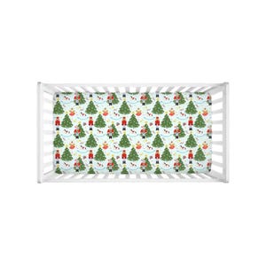 Könnte beinhalten: Ein Babybettlaken mit einem sich wiederholenden Muster aus Weihnachtsbäumen, Spielzeugsoldaten und Pferden auf hellblauem Hintergrund. Das Design umfasst rote, grüne und goldene Akzente und schafft ein festliches Urlaubsthema.