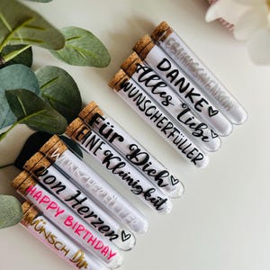 May include: Clear glass tubes with cork stoppers, filled with a white substance. Each tube has black or gold text, including phrases like "Happy Birthday" and "Thank You." The tubes are arranged on a white surface, with green leaves and white flowers in the background.