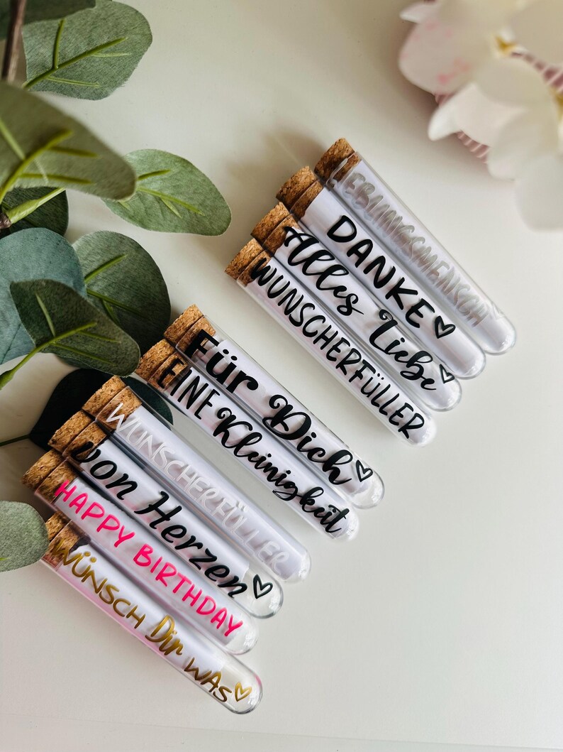 May include: Clear glass tubes with cork stoppers, filled with a white substance. Each tube has black or gold text, including phrases like "Happy Birthday" and "Thank You." The tubes are arranged on a white surface, with green leaves and white flowers in the background.