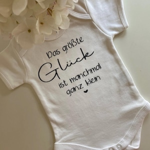 May include: White baby onesie with black text that reads "Das größte Glück ist manchmal ganz klein" with a small heart symbol.