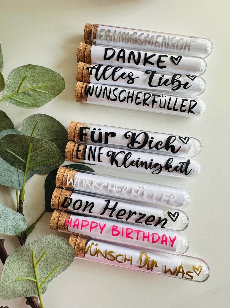 May include: A collection of glass tubes with cork stoppers, each containing a rolled-up message. The tubes are labeled with various phrases in German, including "LIEBLINGSMENSCH", "DANKE", and "HAPPY BIRTHDAY". The background includes green foliage.