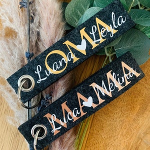 May include: Two black felt keychains with gold and rose gold lettering. One keychain says "OMA Luana Leyla" with a gold heart and the other says "MAMA Luca Melina" with a rose gold heart.