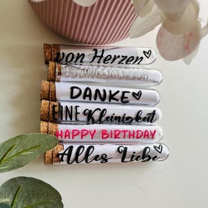 May include: A stack of glass tubes with cork stoppers, each containing a rolled-up message. The messages are written in black and pink script, including "von Herzen", "LIEBLINGSMENSCH", "DANKE", "EINE Kleinigkeit", "HAPPY BIRTHDAY", and "Alles Liebe".